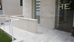Fachada moderna de un edificio con entrada de vidrio, pared color beige y un pequeño área de estar exterior con barandillas blancas.