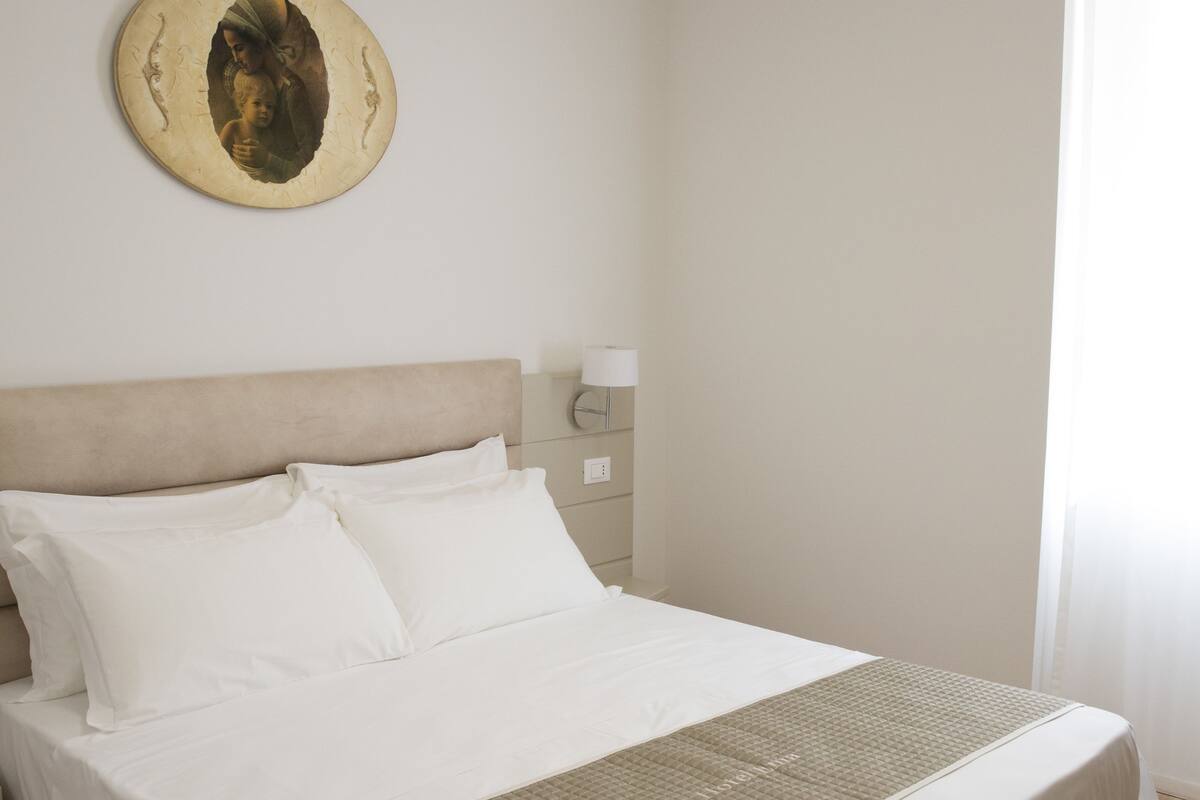 A neatly made bed with white linens and a beige headboard, a bedside table with a lamp, and a wall-mounted decorative plate.