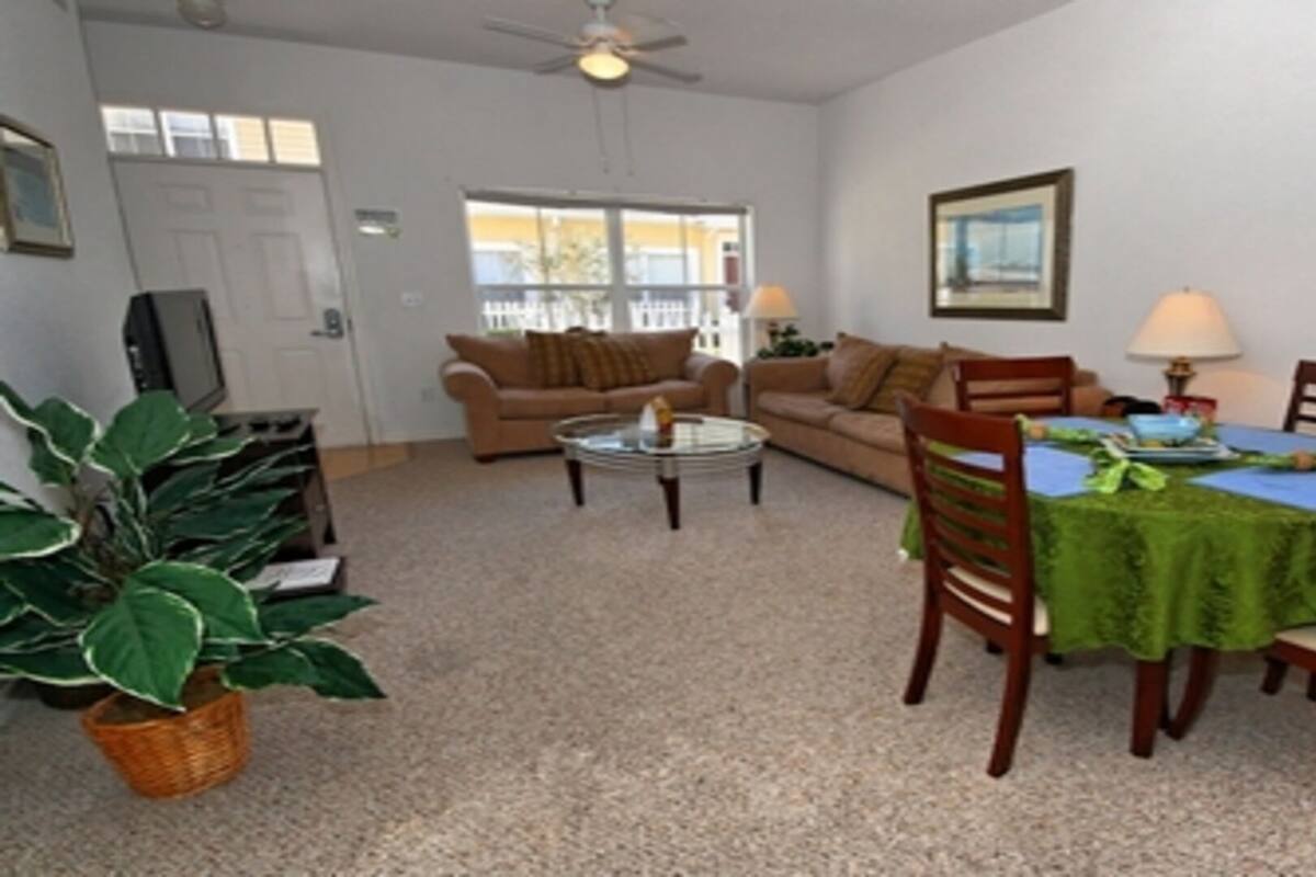 A living room with a sofa, a coffee table, and a dining table with chairs. There is a plant in a basket and a ceiling fan.