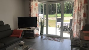 A modern living room with a flat-screen TV, a sofa, a coffee table, and a sliding glass door leading to an outdoor area with a table and chairs. - Ferienwohnung Zweisamkeit (Gelbensande)