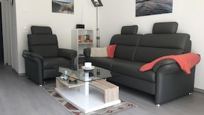 A modern living room with a glass coffee table, two black leather sofas, a bookshelf, and two wall-mounted speakers. - Ferienwohnung Zweisamkeit (Gelbensande)