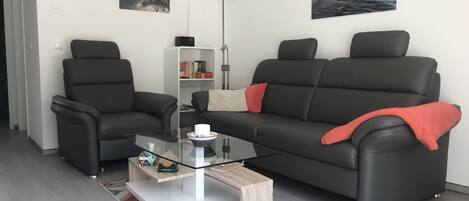 Ein modernes Wohnzimmer mit einem Glastisch, zwei schwarzen Ledersofas, einem Bücherregal und zwei Wandlautsprechern.