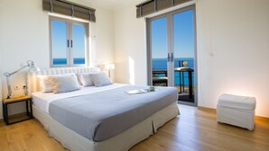 Una camera da letto con un letto grande, un comodino con una lampada, un tavolino con una sedia e vista sul mare tramite una porta scorrevole.