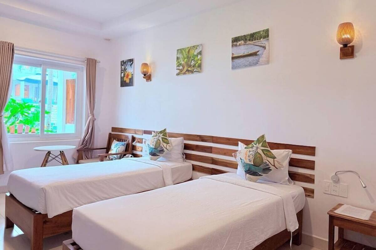 Une chambre d’hôtel avec deux lits, une petite table, une chaise, une fenêtre avec des rideaux et des œuvres d’art accrochées au mur.