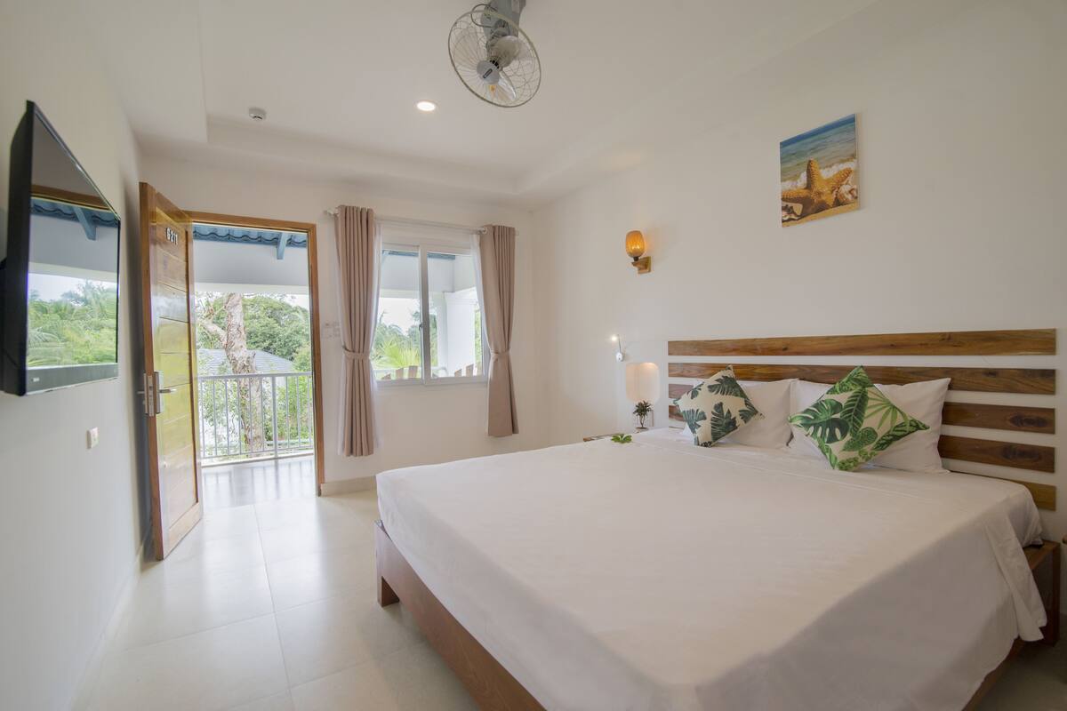 Une chambre avec un grand lit, un ventilateur de plafond, une fenêtre avec des rideaux, un téléviseur fixé au mur et un tableau représentant une scène de plage.
