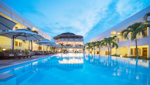 A swimming pool with lounge chairs and umbrellas, surrounded by buildings with balconies and palm trees. - Palma Resort Phu Quoc, Trademark Collection by Wyndham (Phu Quoc)