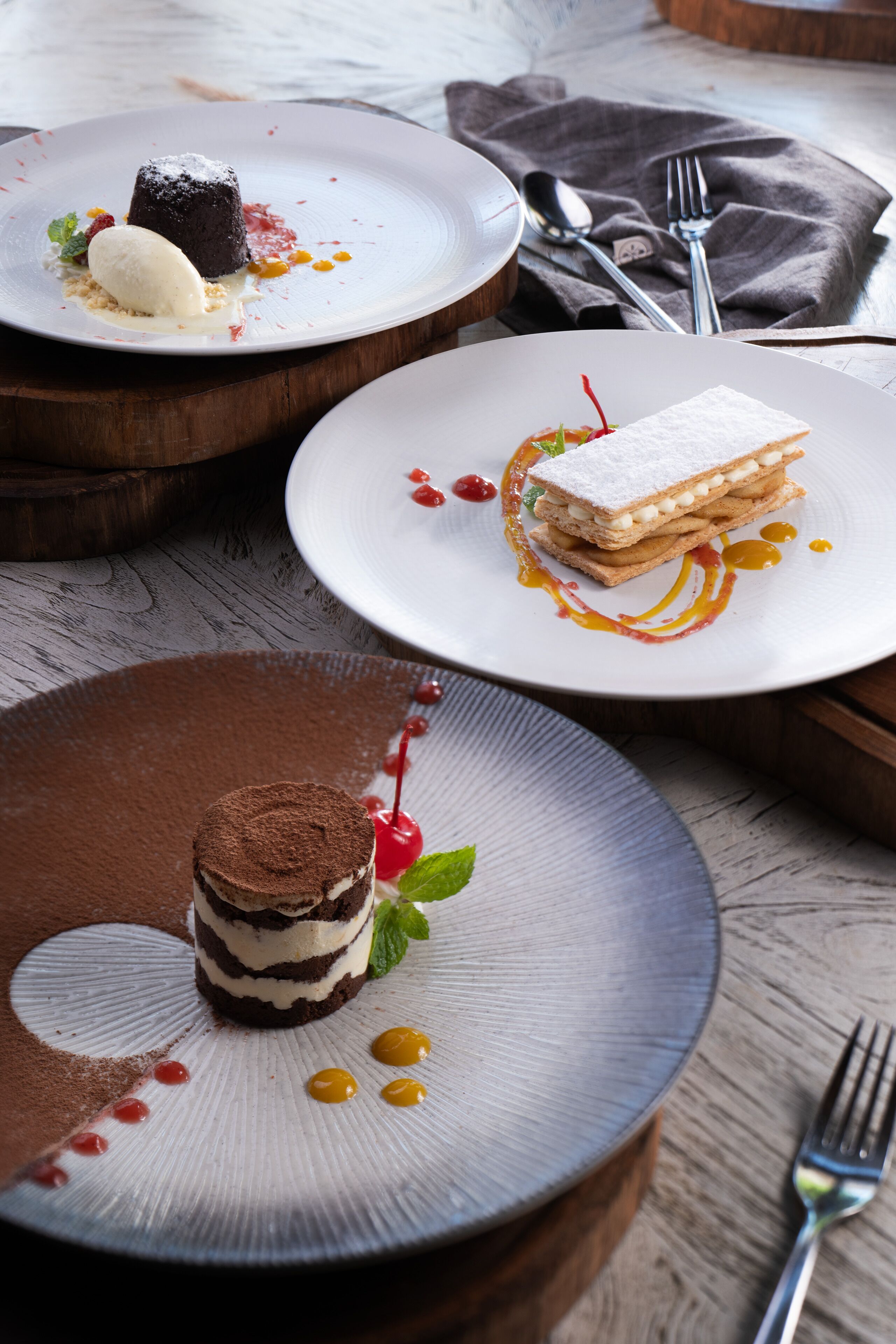 Trois assiettes à dessert, sur lesquelles sont disposés différents desserts, sont posées sur une table en bois.