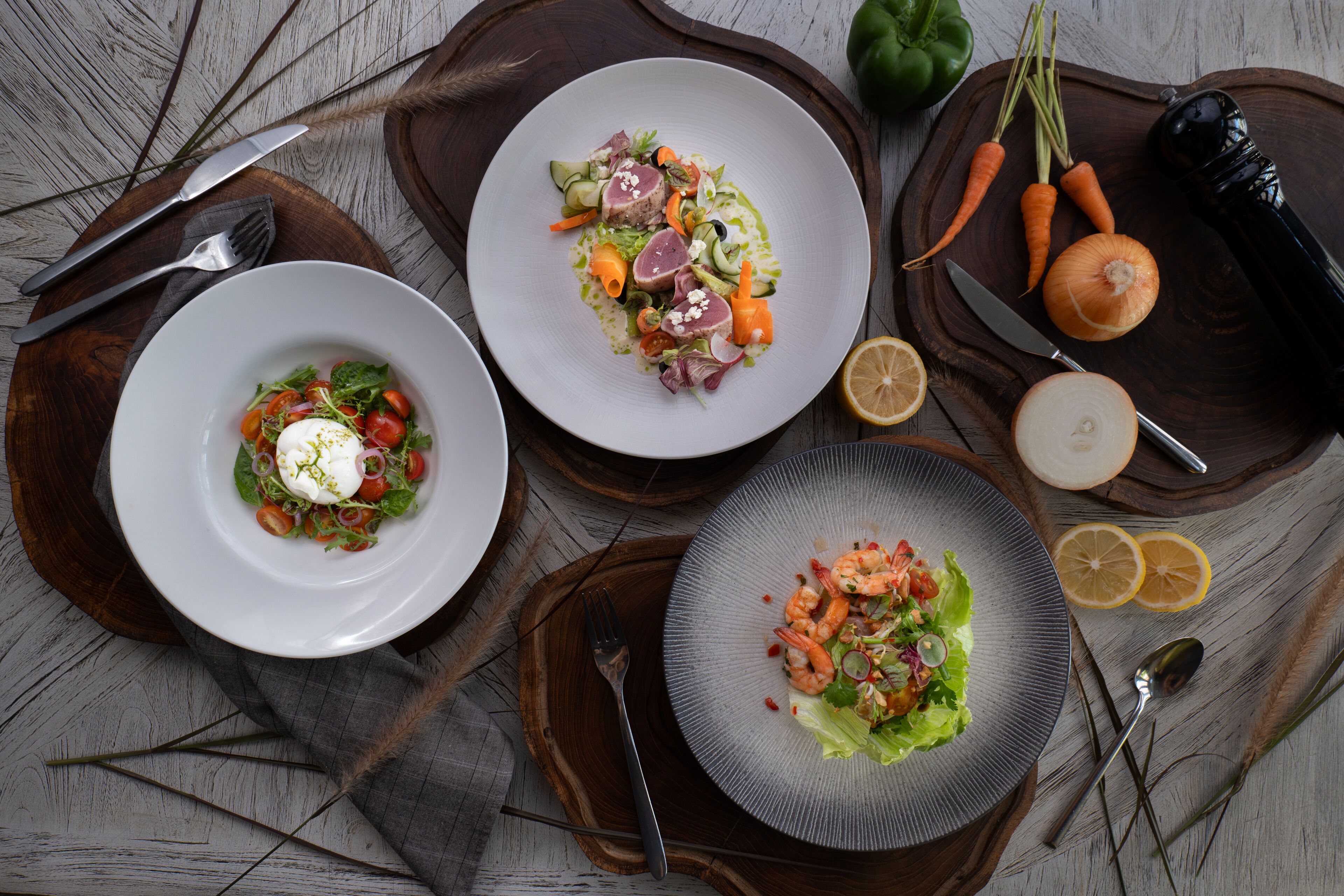 Three plates of food on wooden serving boards with a knife, fork, and lemon slices nearby.