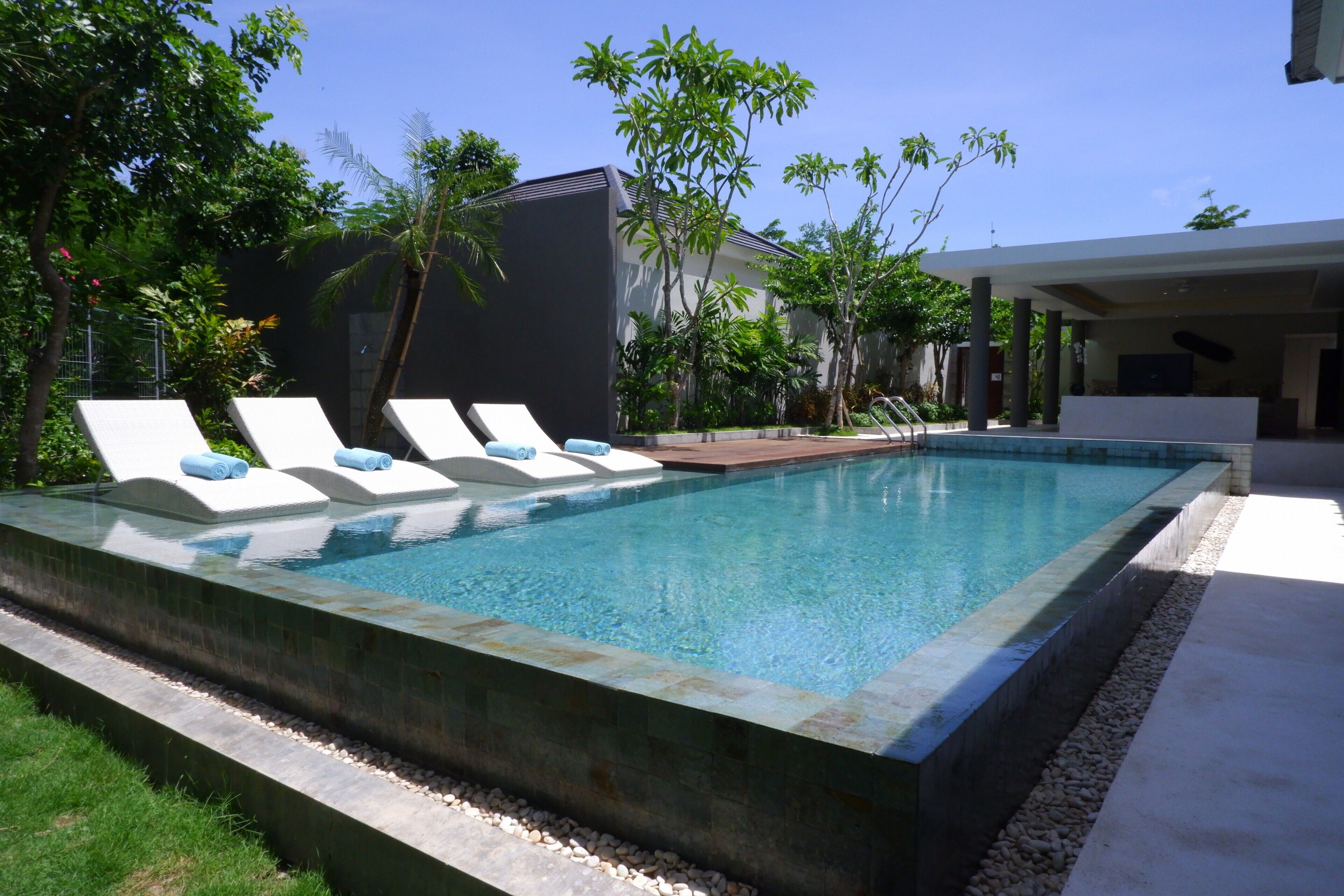 Piscina rectangular con sillones de descanso y una zona cubierta, rodeada de vegetación y un edificio moderno.
