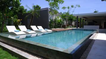 Piscina rectangular con sillones de descanso y una zona cubierta, rodeada de vegetación y un edificio moderno.