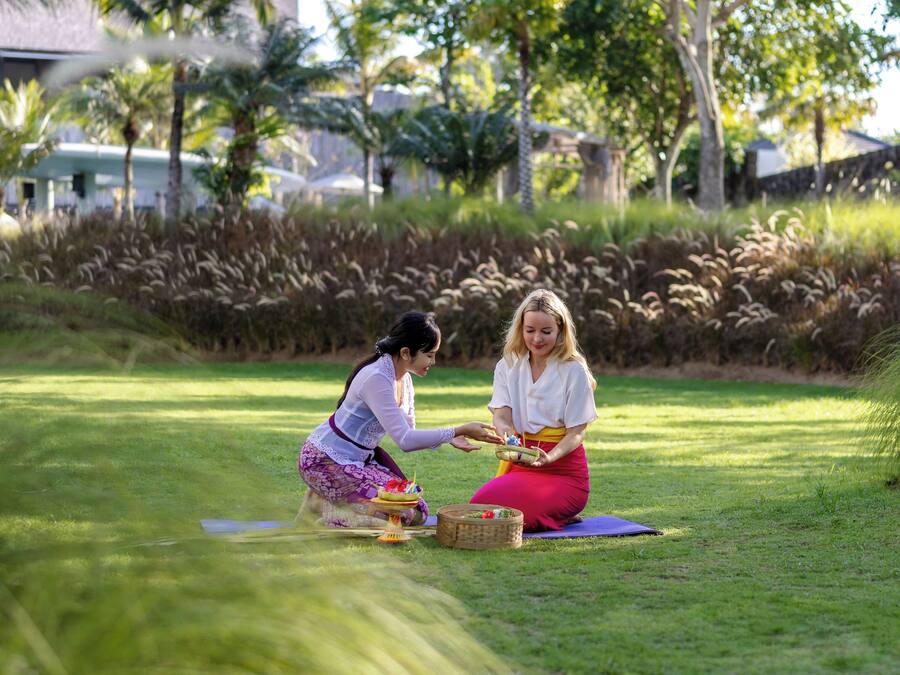 Two people sitting on a grassy area with a picnic setup, surrounded by greenery and trees.