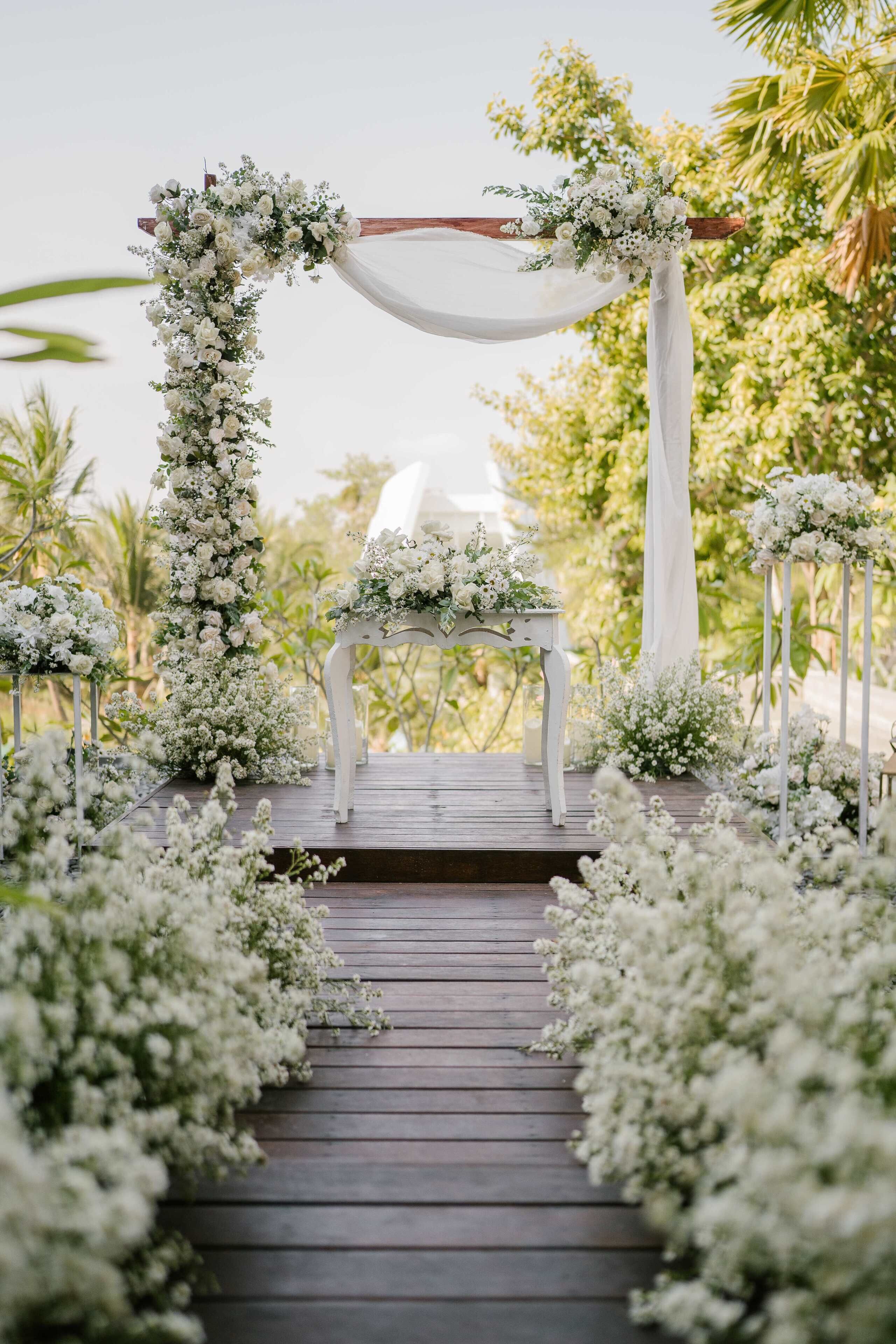 A wooden pathway leading to a white chair under a floral archway.