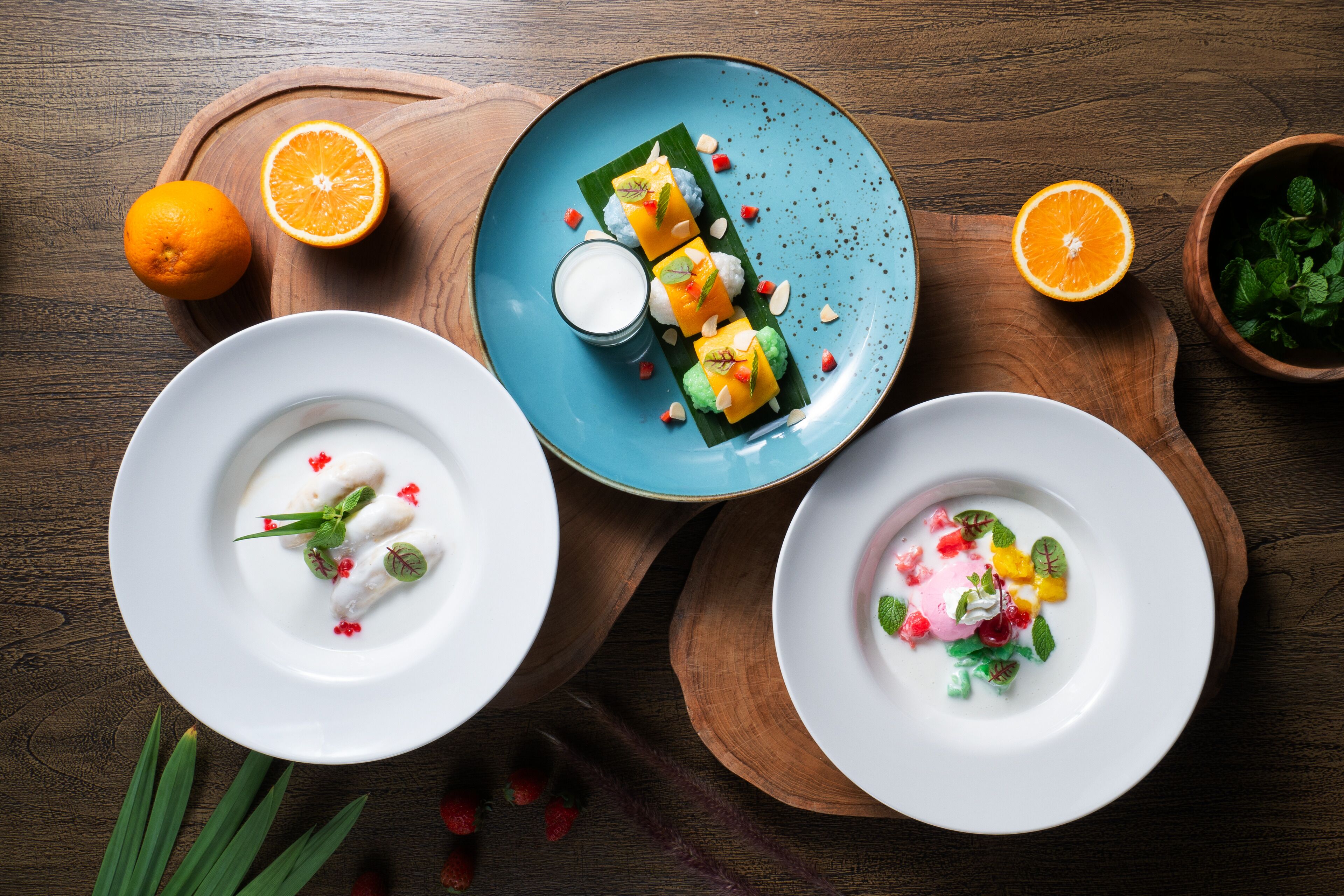 A wooden table with three white plates containing various dishes, a blue plate with a dessert, and two whole oranges.