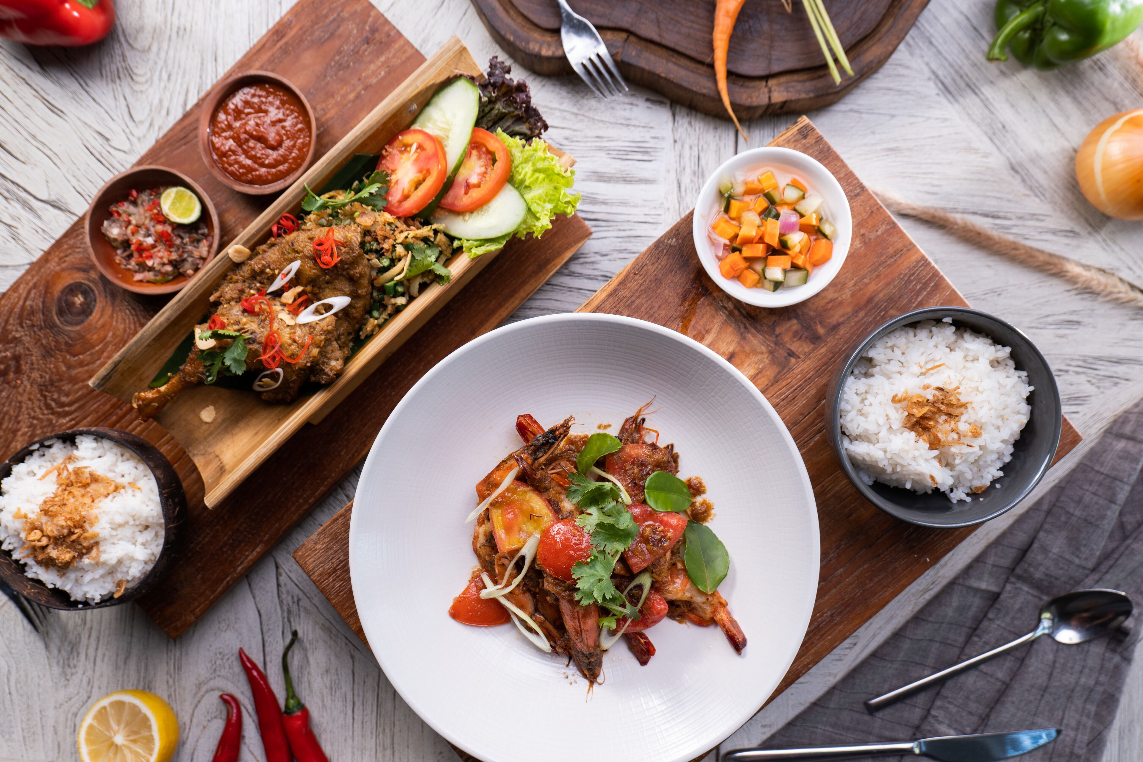 Una mesa de madera con diversos platos, incluyendo un pescado, arroz y verduras, junto con recipientes de condimentos y una tabla de madera con más alimentos.