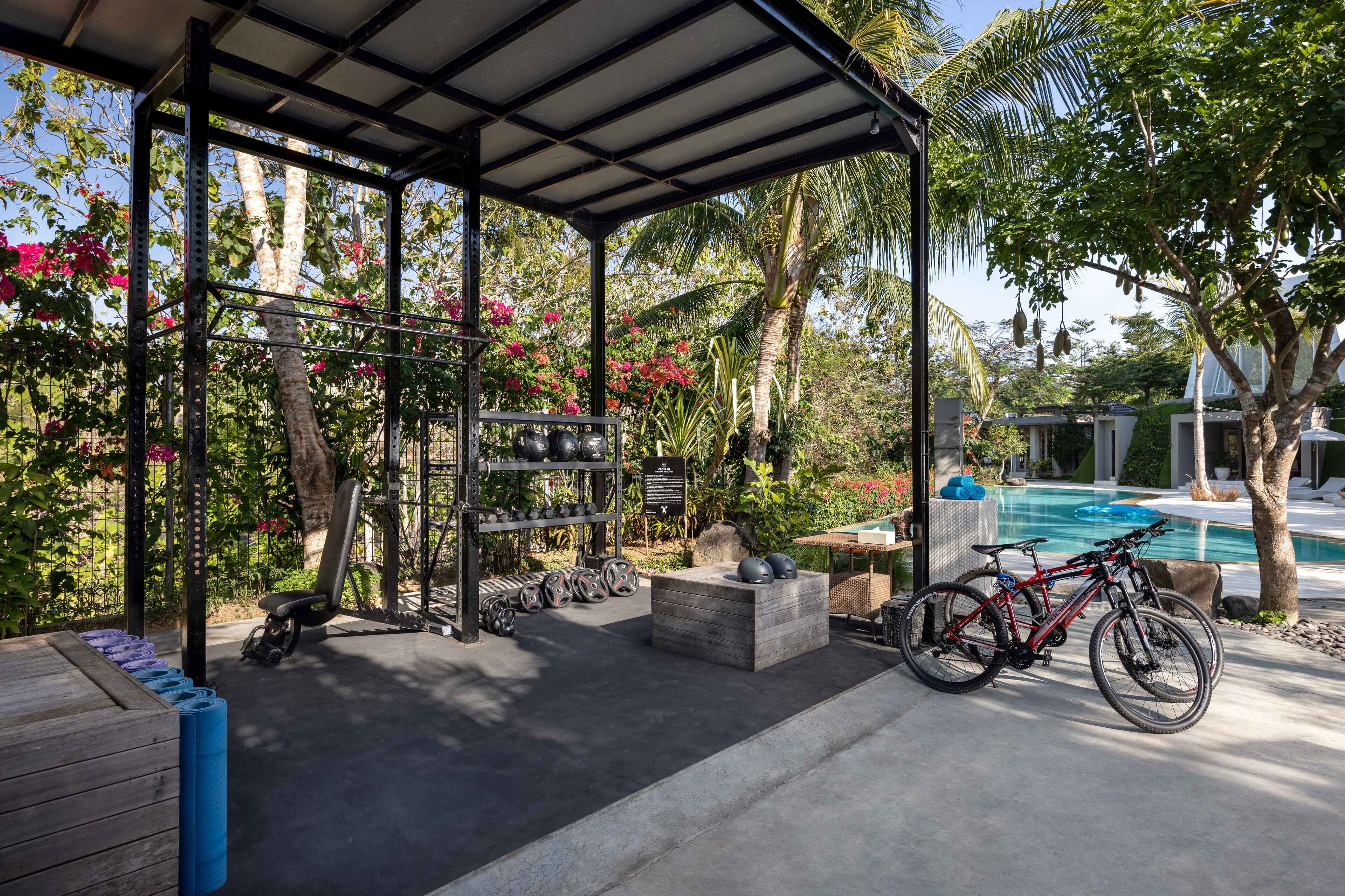 Un espacio al aire libre techado con equipo de ejercicio, una bicicleta y una piscina al fondo.