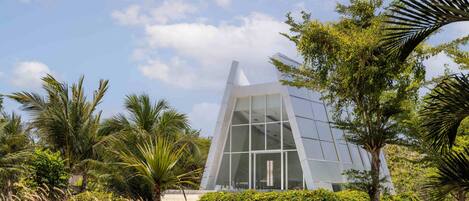 Un edificio moderno con fachada de vidrio geométrica rodeado de exuberante vegetación y palmeras.