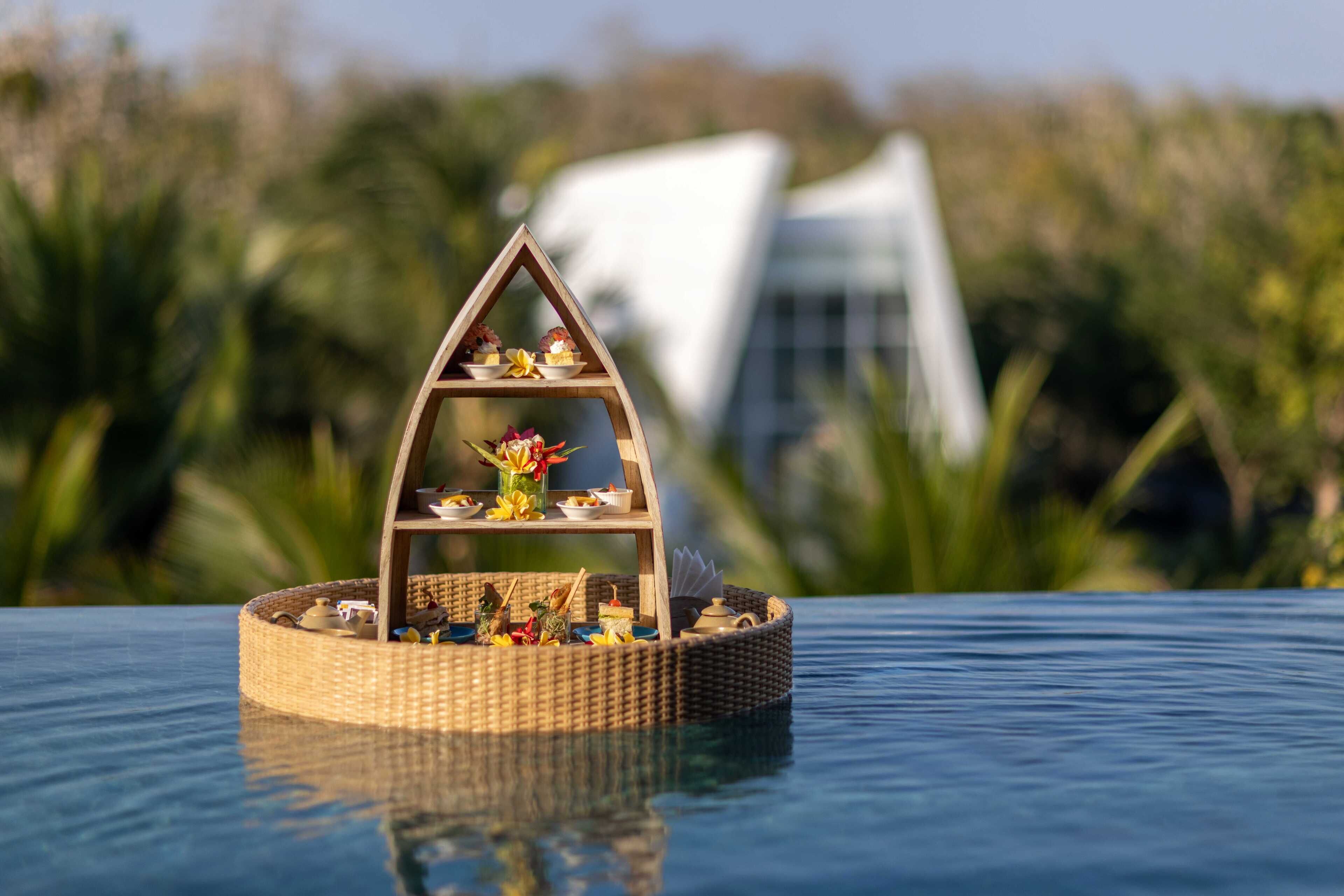 Een drijvend houten schap met diverse objecten, omringd door water, met een modern gebouw op de achtergrond.