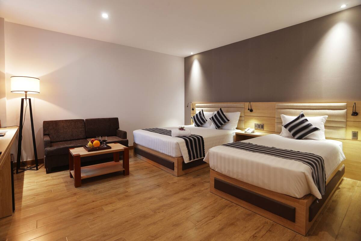 Quarto de hotel com duas camas, um sofá, uma mesa de centro com frutas e uma luminária.
