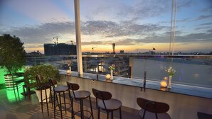 A rooftop bar with bar stools and potted plants, overlooking a cityscape with construction cranes and a sunset.
