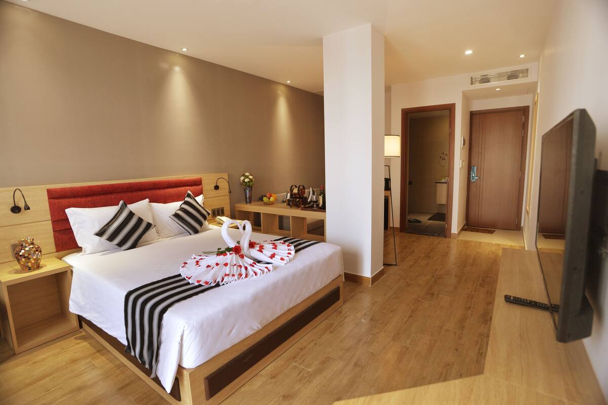 Quarto de hotel com uma cama grande, mesa de cabeceira, televisão e piso de madeira.
