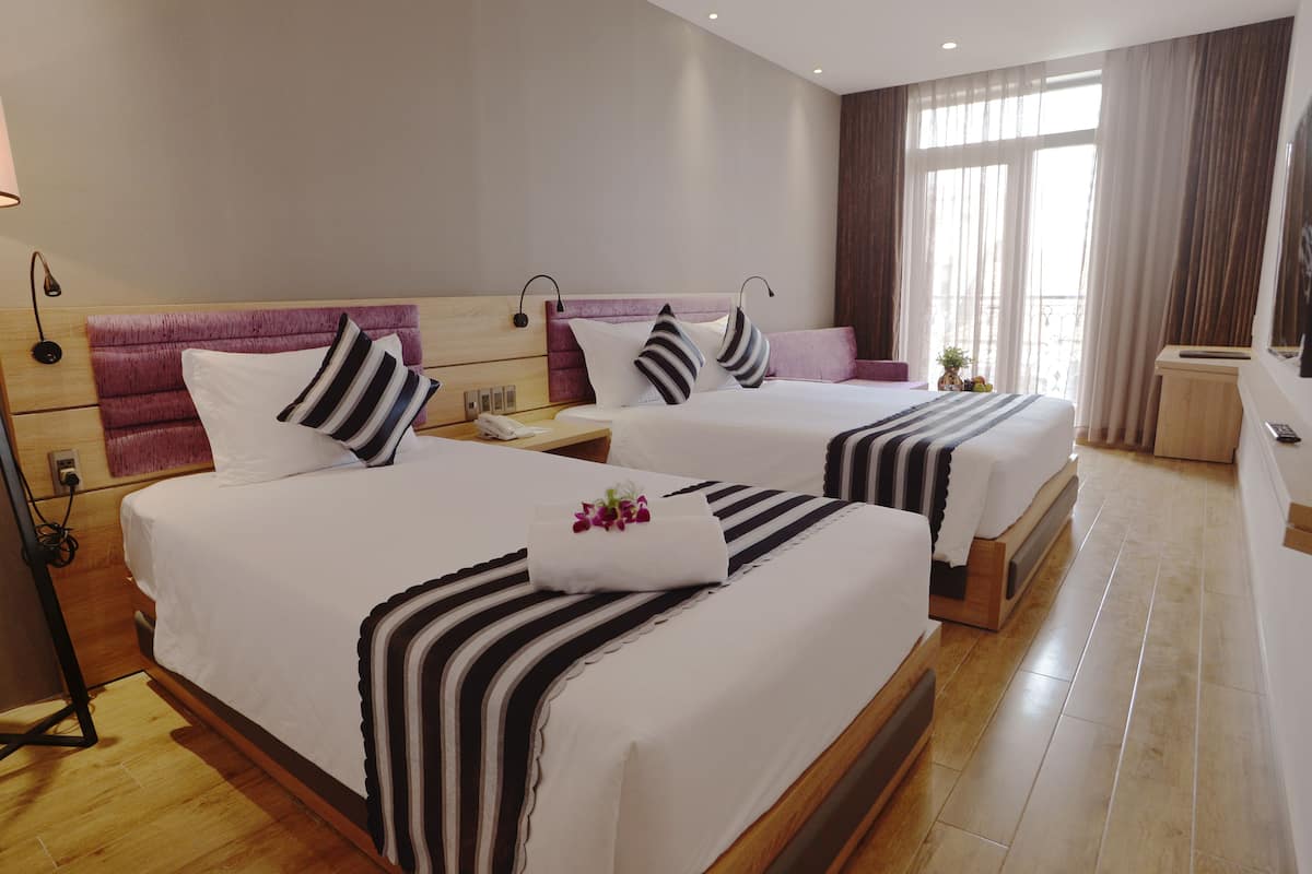 Quarto de hotel com duas camas, cada uma com cabeceira de madeira, roupa de cama branca e almofadas listradas. Em cada cama, um criado-mudo com abajur e telefone.