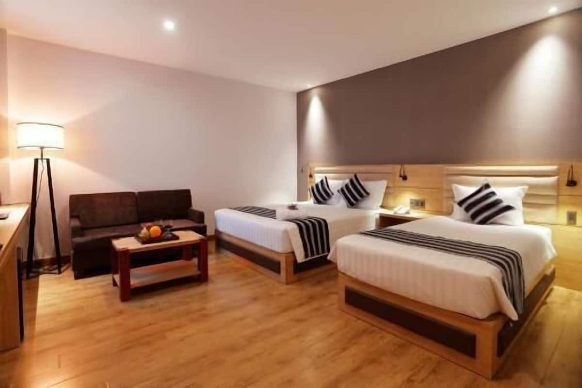 Quarto de hotel com duas camas, uma escrivaninha, uma luminária e uma mesinha com frutas.
