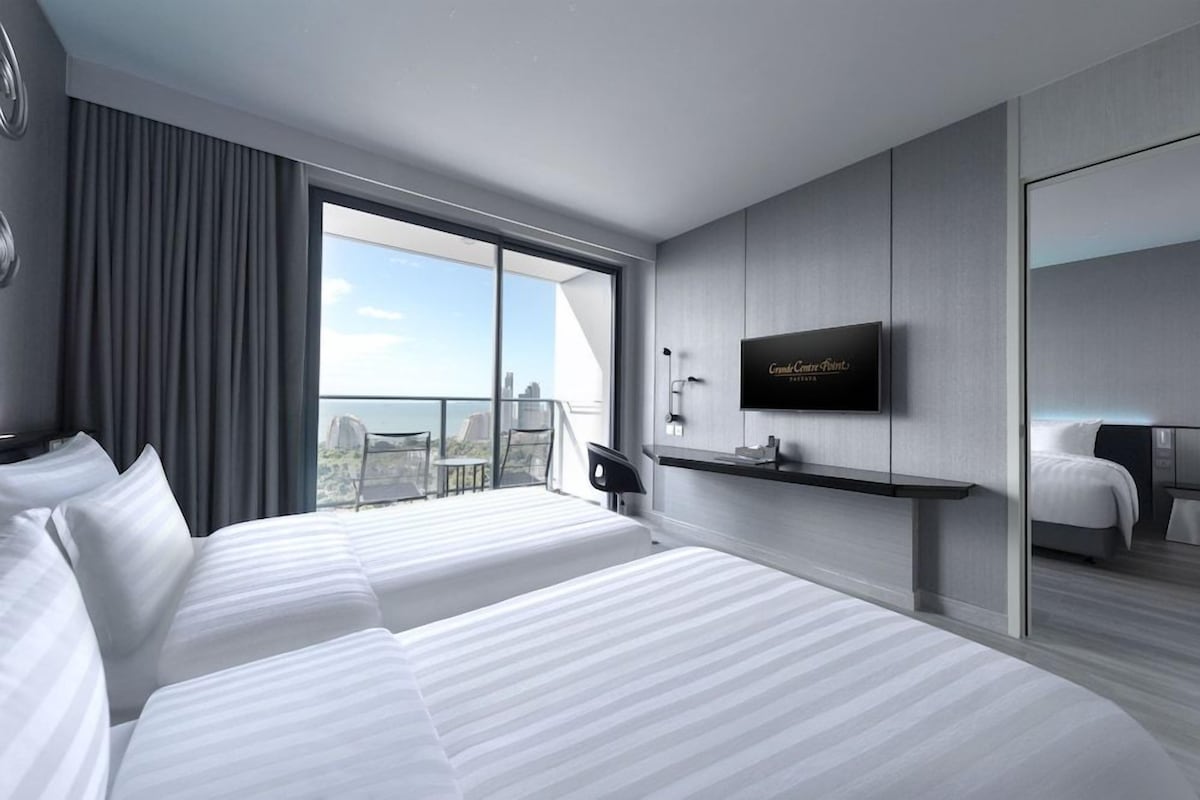 A modern hotel room with two beds, a TV, a balcony with a city view, and a large window.