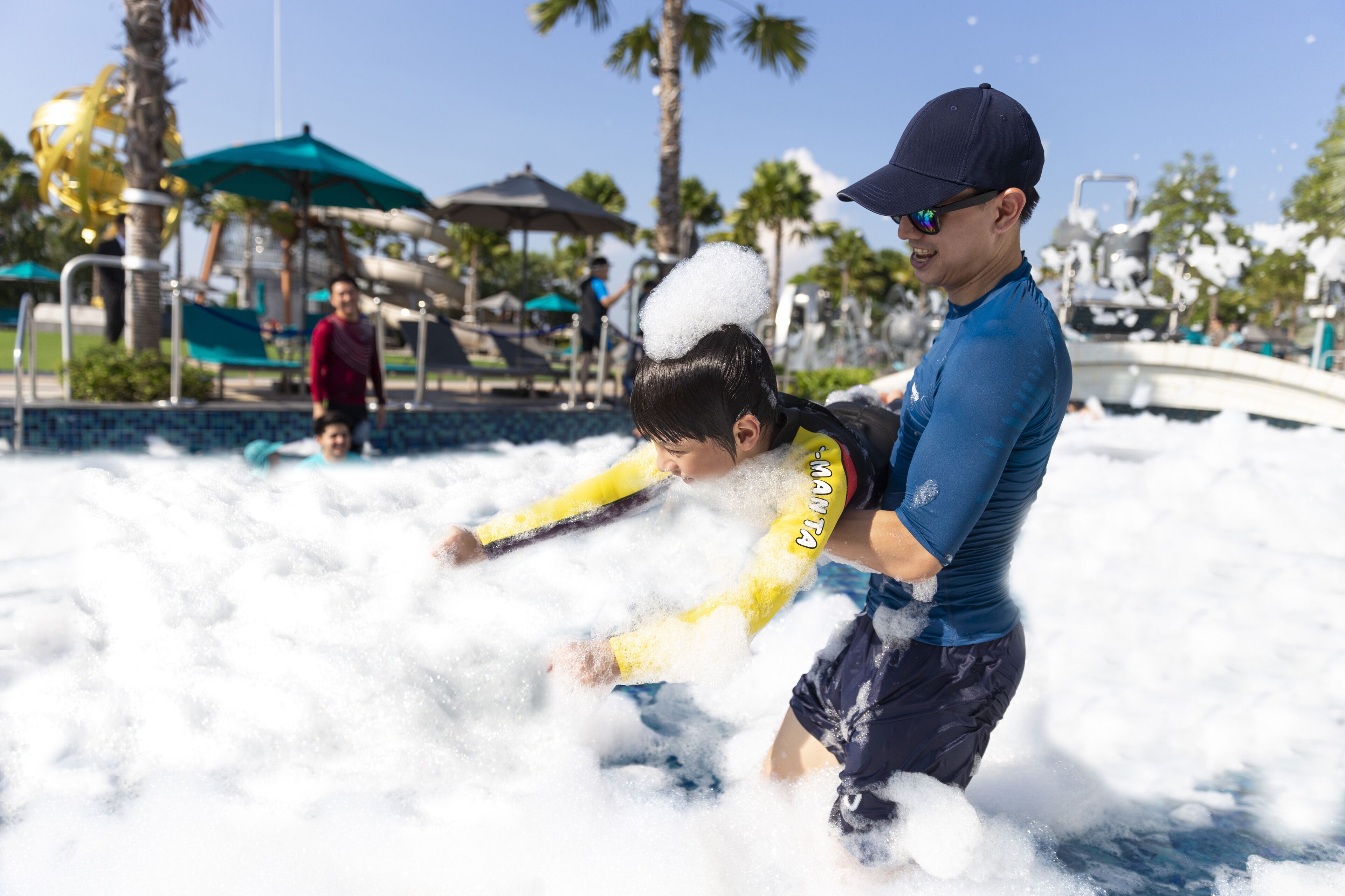 A man and a child playing in a water park with palm trees and a boat in the background.