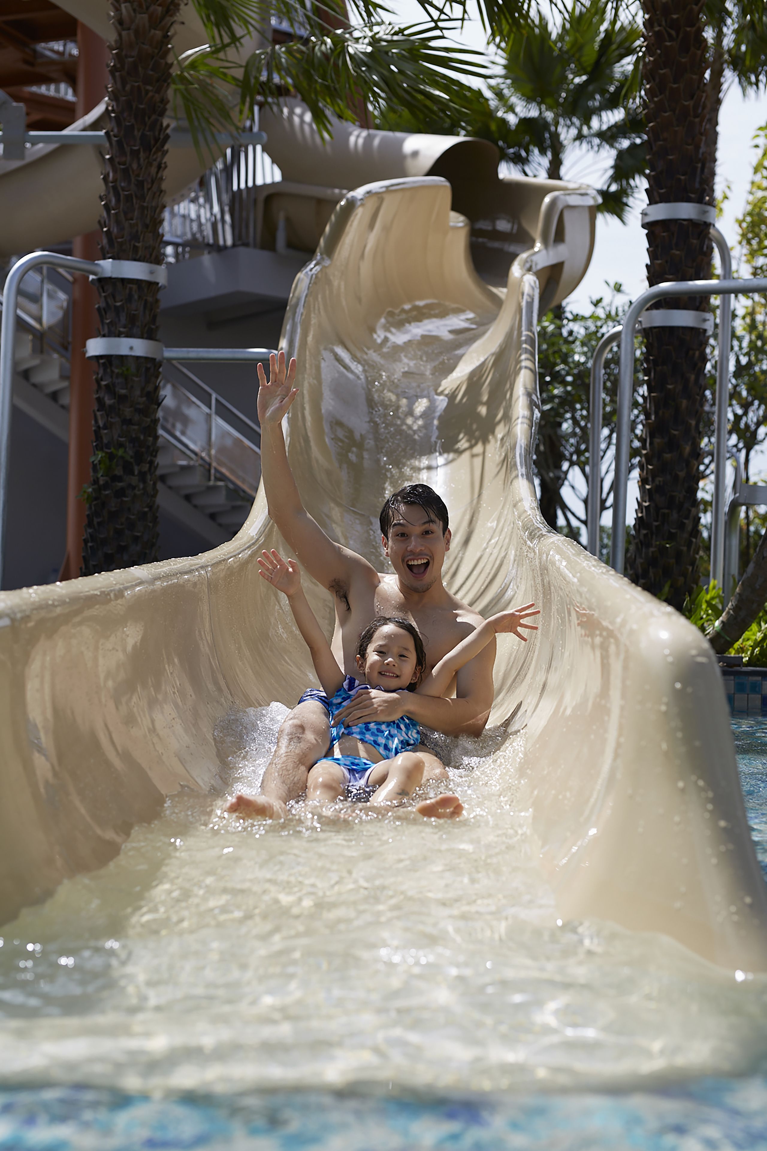 Tobogán de agua en un resort con piscina, palmeras y una escalera de fondo.