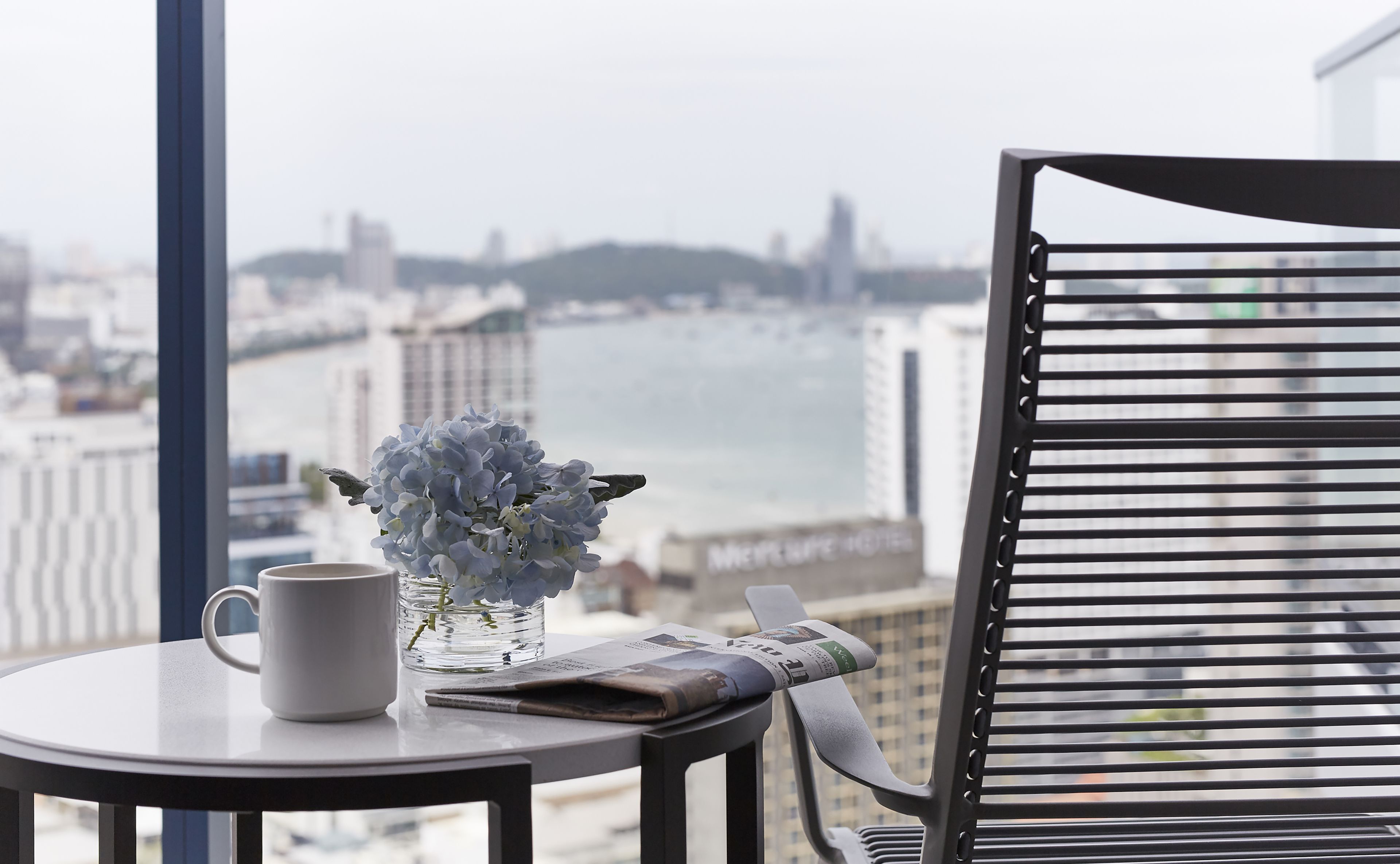 A table with a white mug, a vase with blue flowers, and a newspaper. A black chair is beside the table.