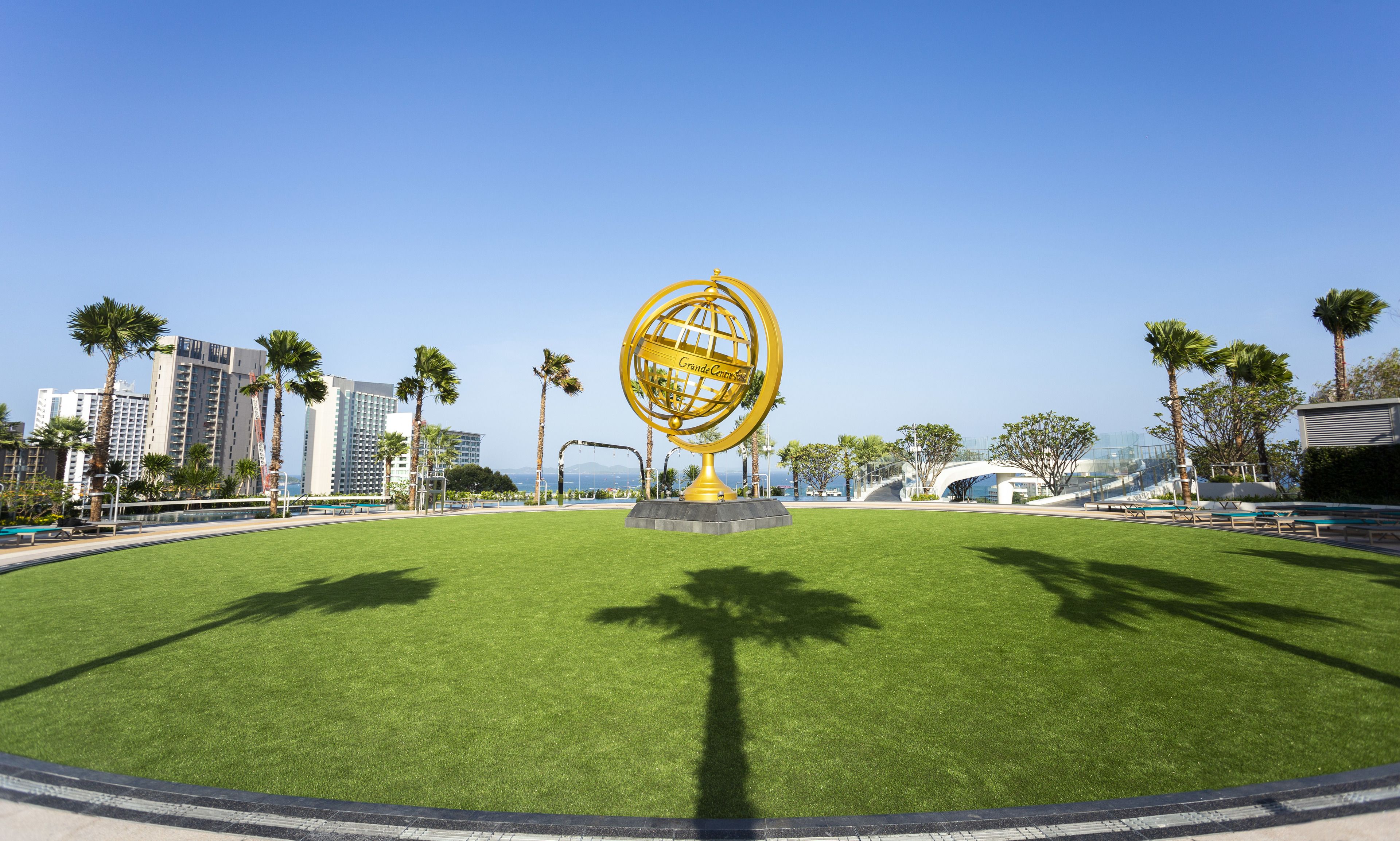 A circular lawn with a golden globe sculpture in the center, surrounded by palm trees and modern buildings in the background.
