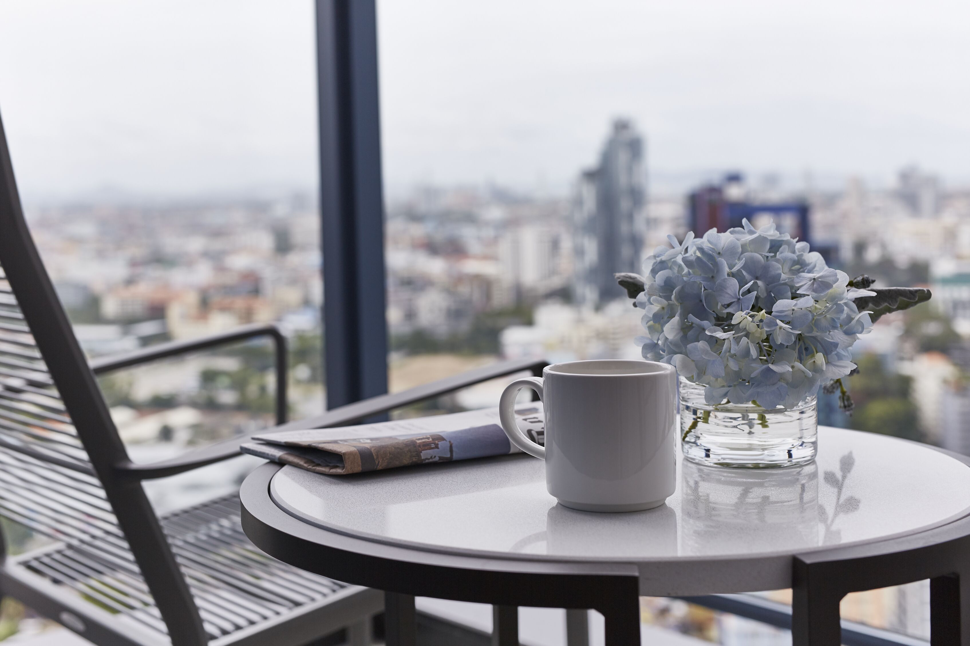 Une table ronde sur laquelle se trouvent une tasse blanche, un vase de fleurs, un journal et, à travers de grandes fenêtres, une vue sur le paysage urbain.