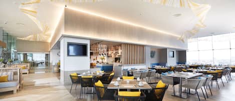 A modern restaurant with a high ceiling and natural light, featuring a mix of seating arrangements and a visible kitchen area.