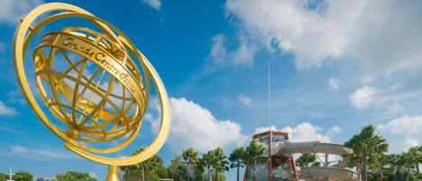 A large golden globe sculpture in a park with a water slide and palm trees.