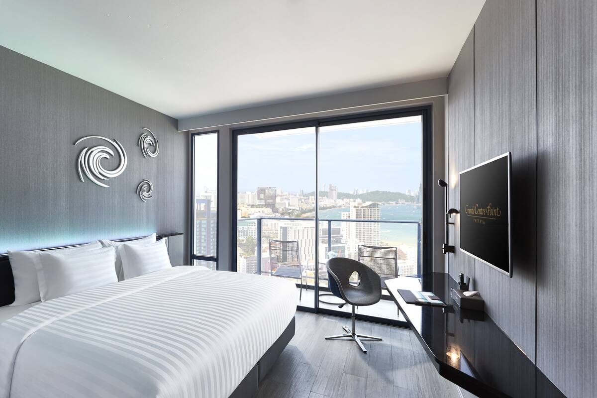 A modern hotel room with a large bed, a desk, a chair, a TV, and a city view through large windows.
