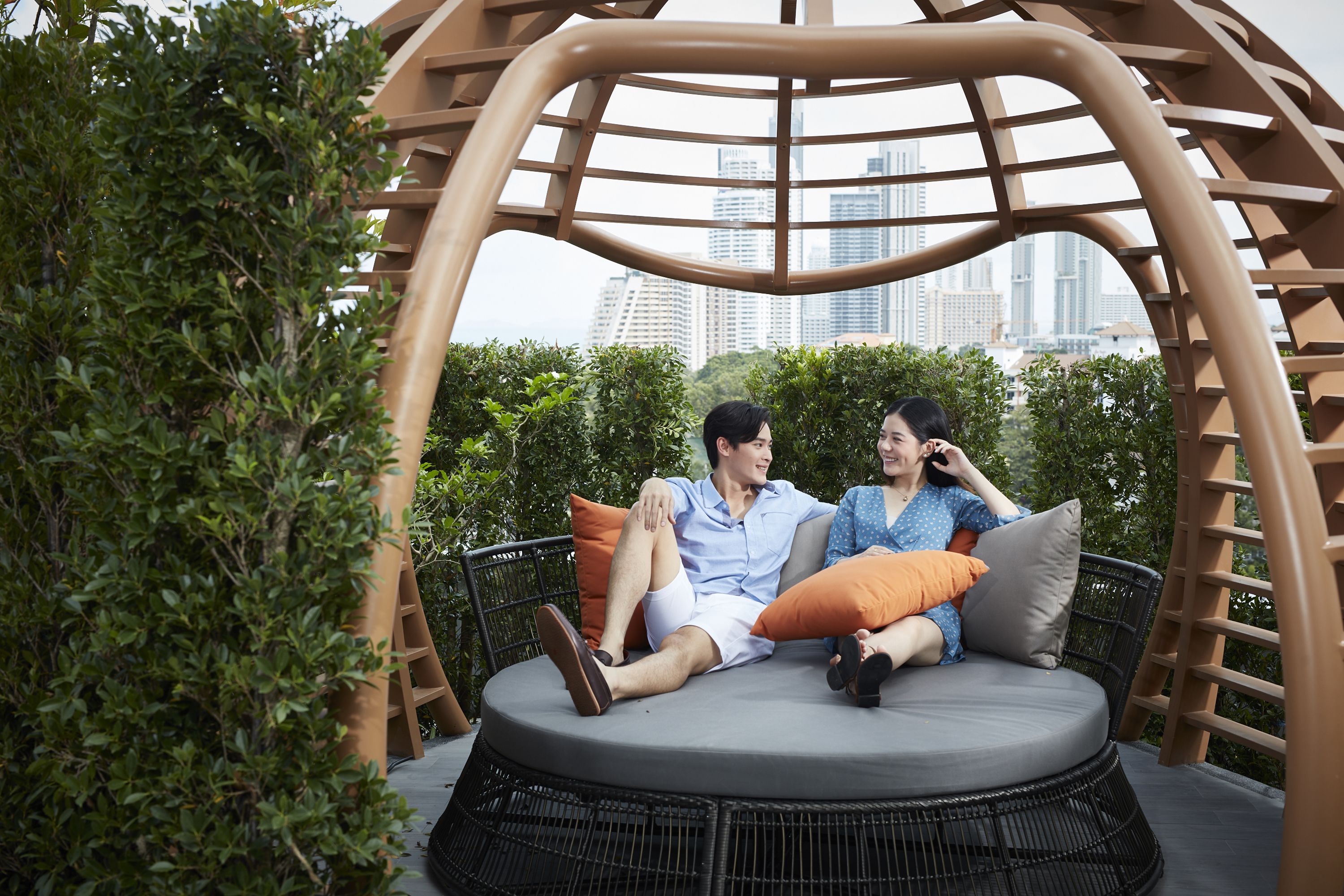 A couple sitting on a round, cushioned outdoor seating area with a cityscape view in the background.