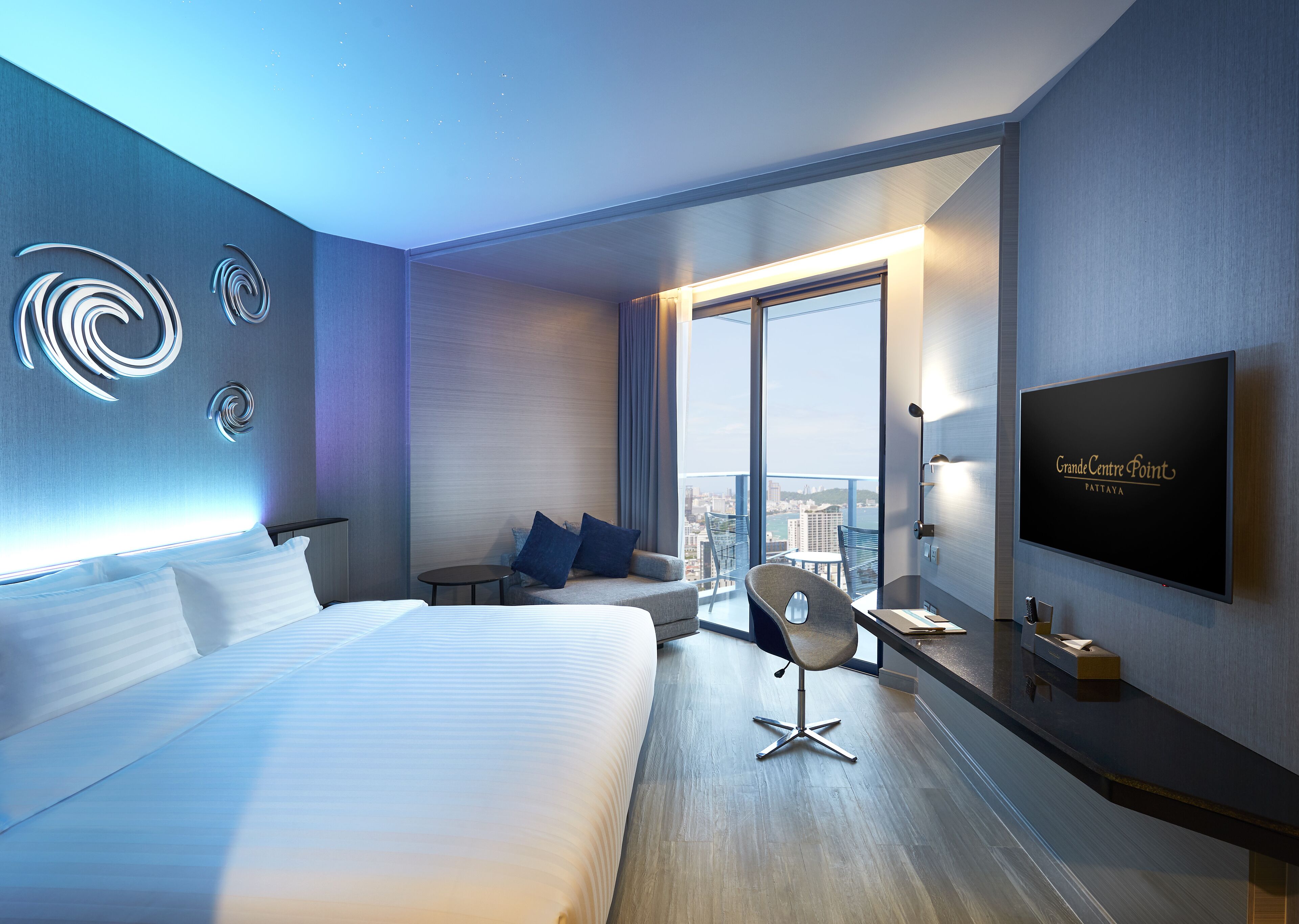 Una habitación de hotel moderna con una cama grande, un escritorio con silla, televisión y vistas a la ciudad a través de amplias ventanas.