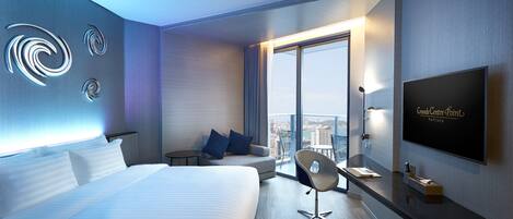 Una habitación de hotel moderna con una cama grande, un escritorio con silla, televisión y vistas a la ciudad a través de amplias ventanas.