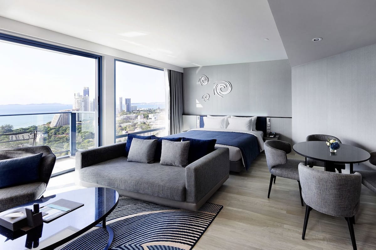 A modern hotel room with a large bed, a seating area with sofas and chairs, a small table, and a city view.