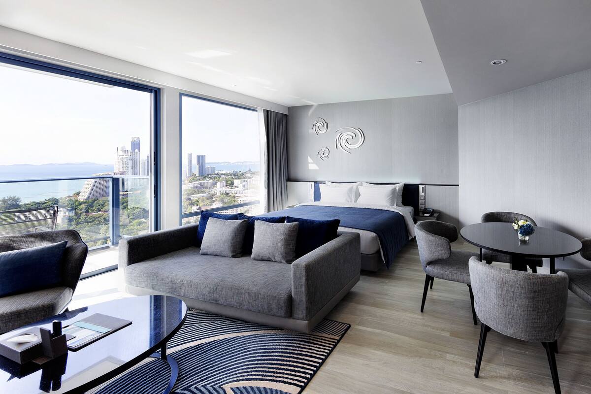 A modern hotel room with a balcony, a bed, a sofa, a dining table, and two chairs.