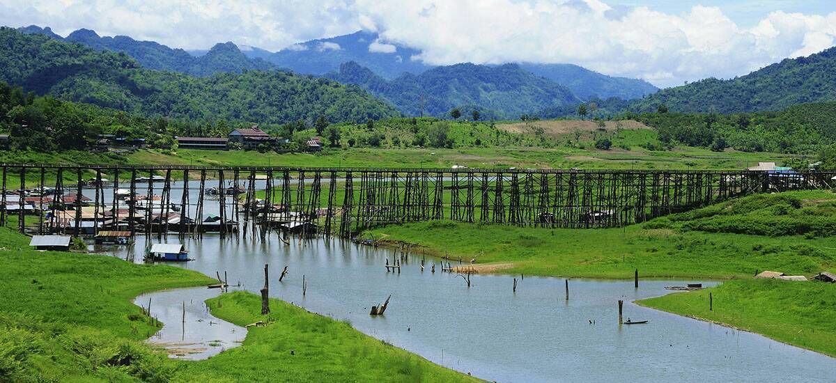 A river with a wooden bridge and boats, surrounded by lush greenery and mountains.