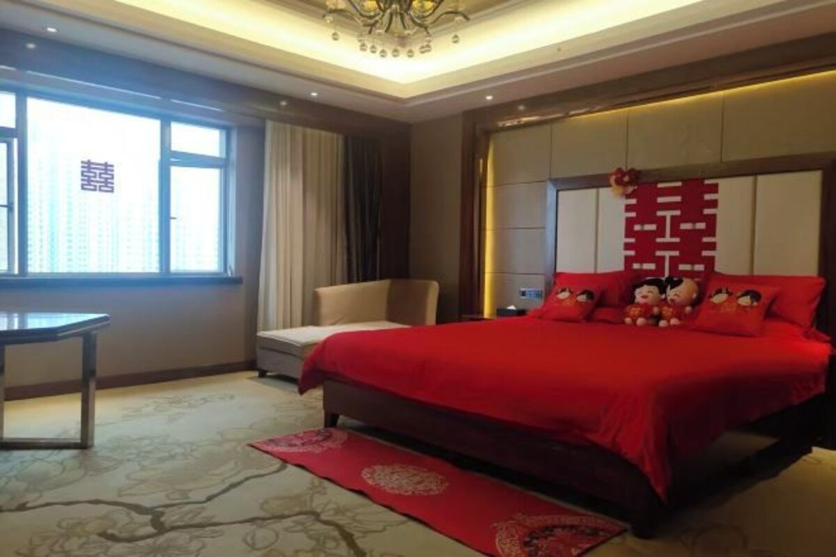 A hotel room with a large bed, a desk, and a chair. There is a window with curtains, a chandelier, and a red decorative pillow on the bed.