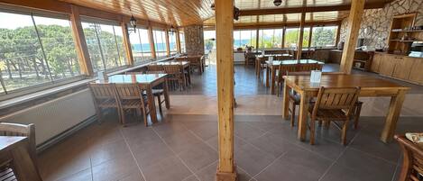 Uma sala de jantar ampla com mesas e cadeiras de madeira, grandes janelas com vista para o exterior e uma parede de destaque em pedra.