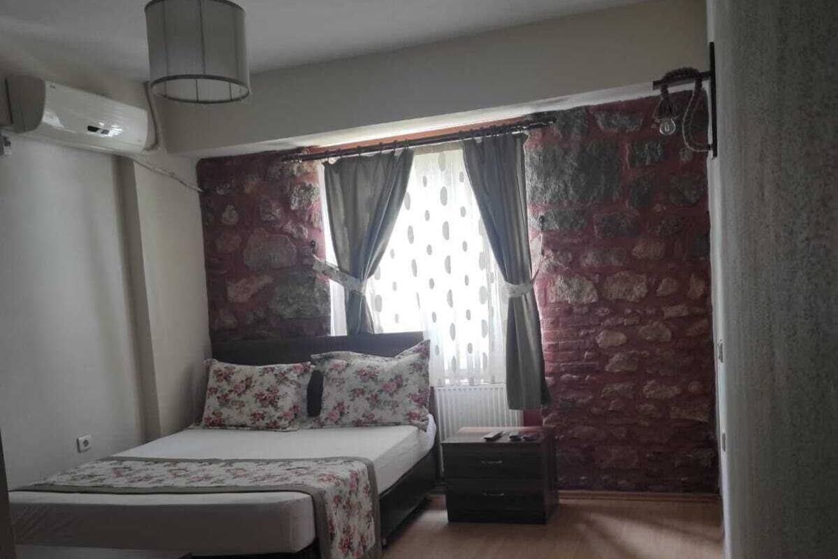 A bedroom with a bed, a nightstand, a dresser, a window with curtains, and a stone wall.