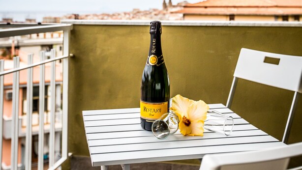 A bottle of Rotari wine, a glass, and a yellow flower on a white table on a balcony with a cityscape view. - B&B Pepito (Cefalù)