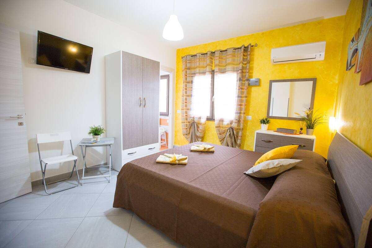 Habitación de hotel con cama, escritorio, silla, armario, televisor, ventana con cortinas y unidad de aire acondicionado.