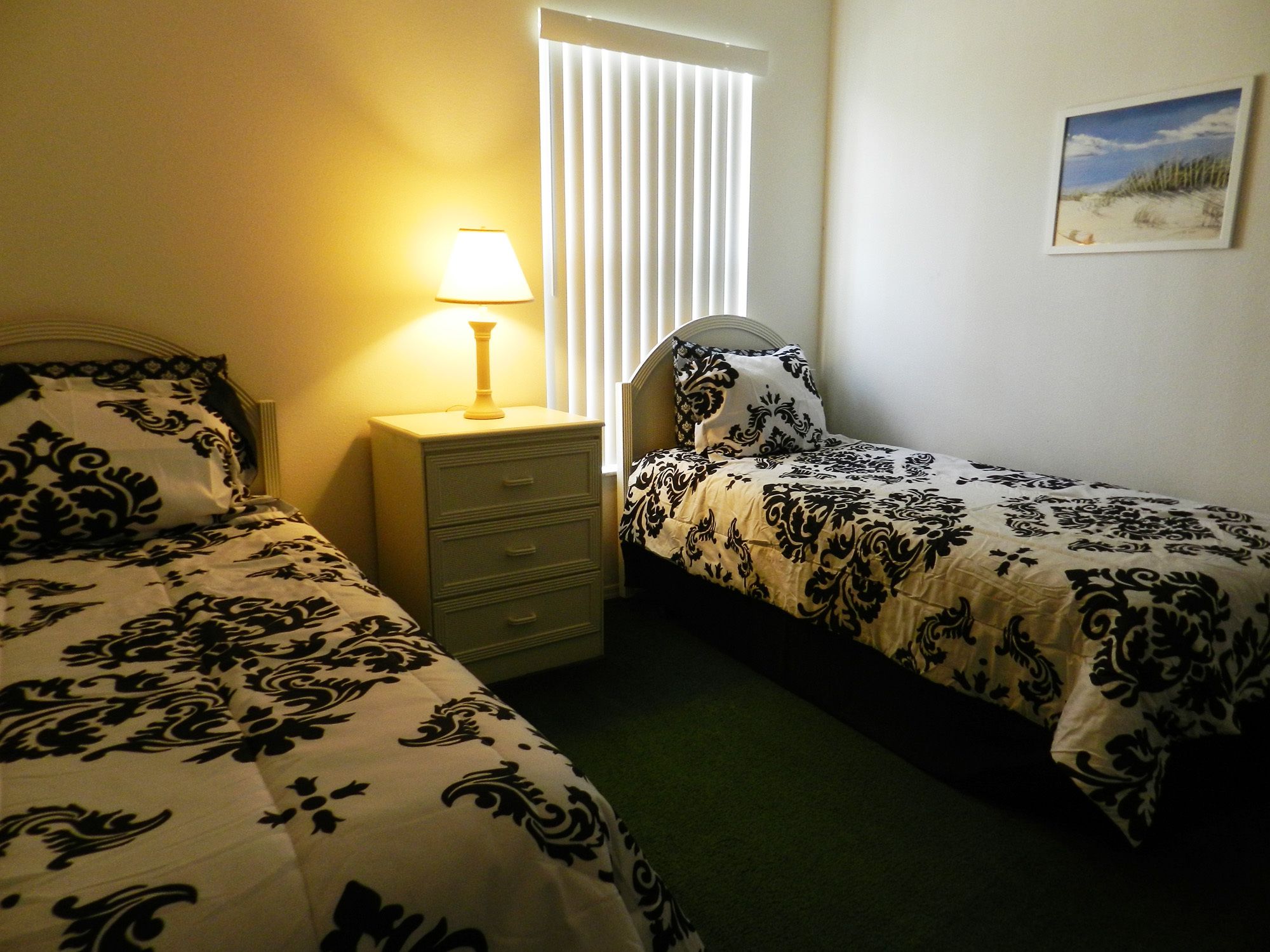 A bedroom with two beds, a nightstand, a lamp, and a window with blinds.