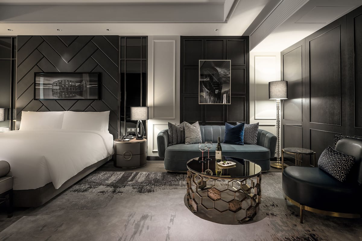A modern hotel room with a large bed, a sofa, a coffee table, and a desk with a lamp.