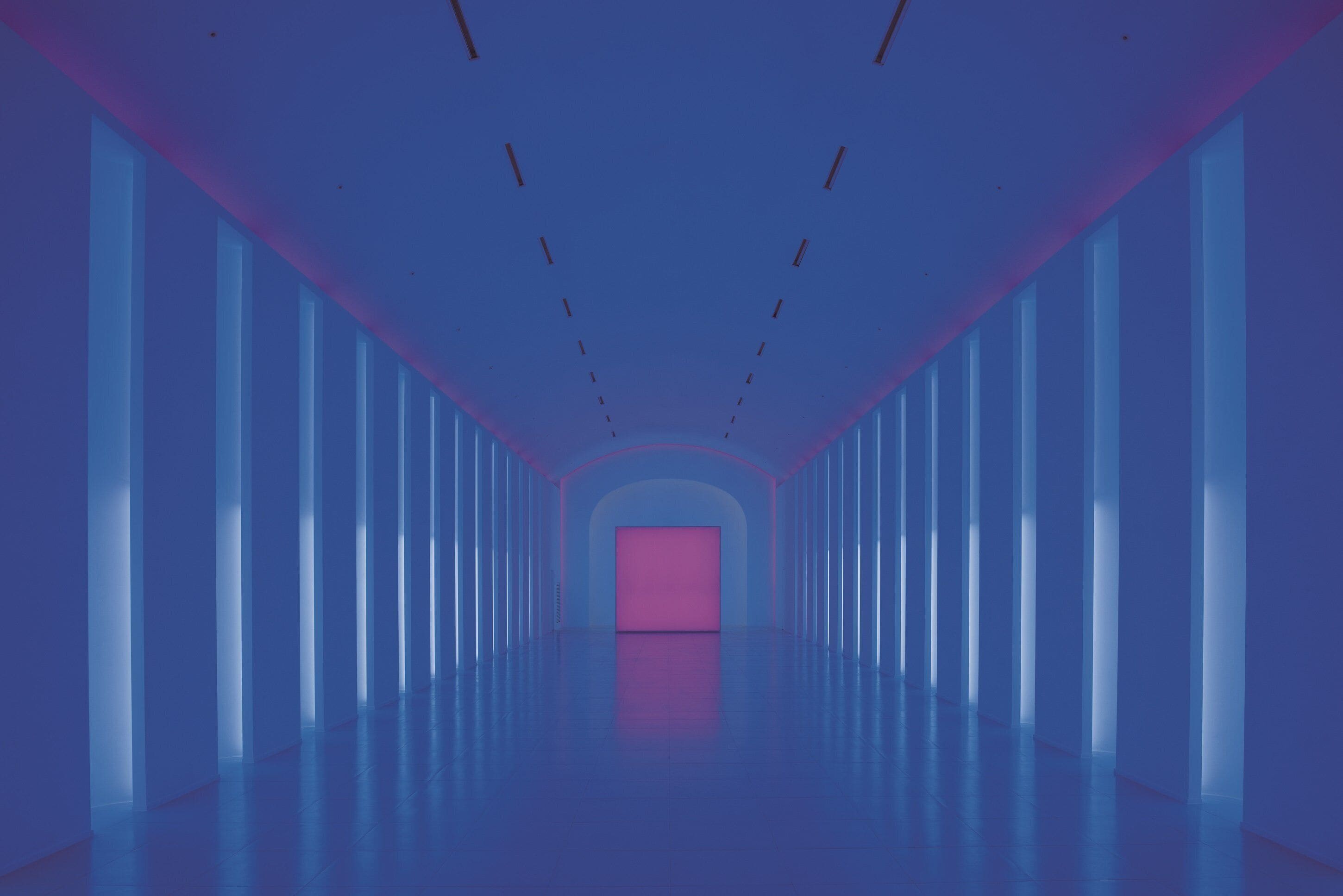 A long, narrow hallway with a central pink door, flanked by tall windows on both sides.