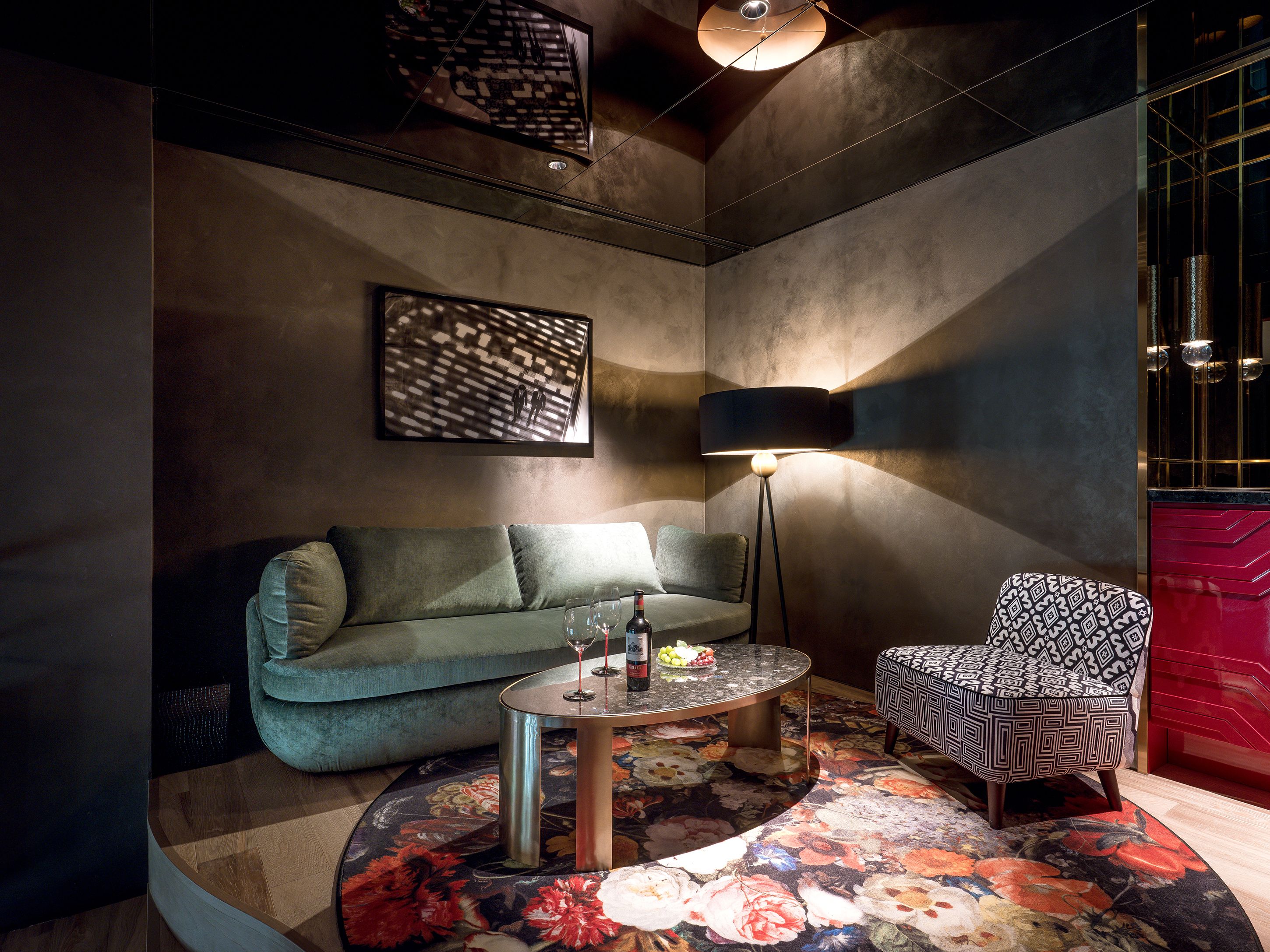 A modern lounge area with a sofa, a patterned armchair, a round table with a floral rug, and a floor lamp.